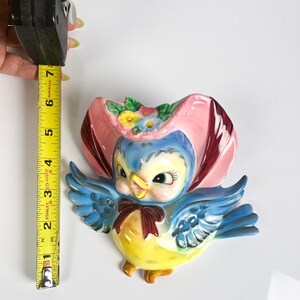 May include: A ceramic wall pocket shaped like a blue bird wearing a pink bonnet with flowers. The bird has a yellow chest and a dark red bow tie. A yellow Stanley measuring tape shows the bird is approximately 7 inches tall from top to bottom.