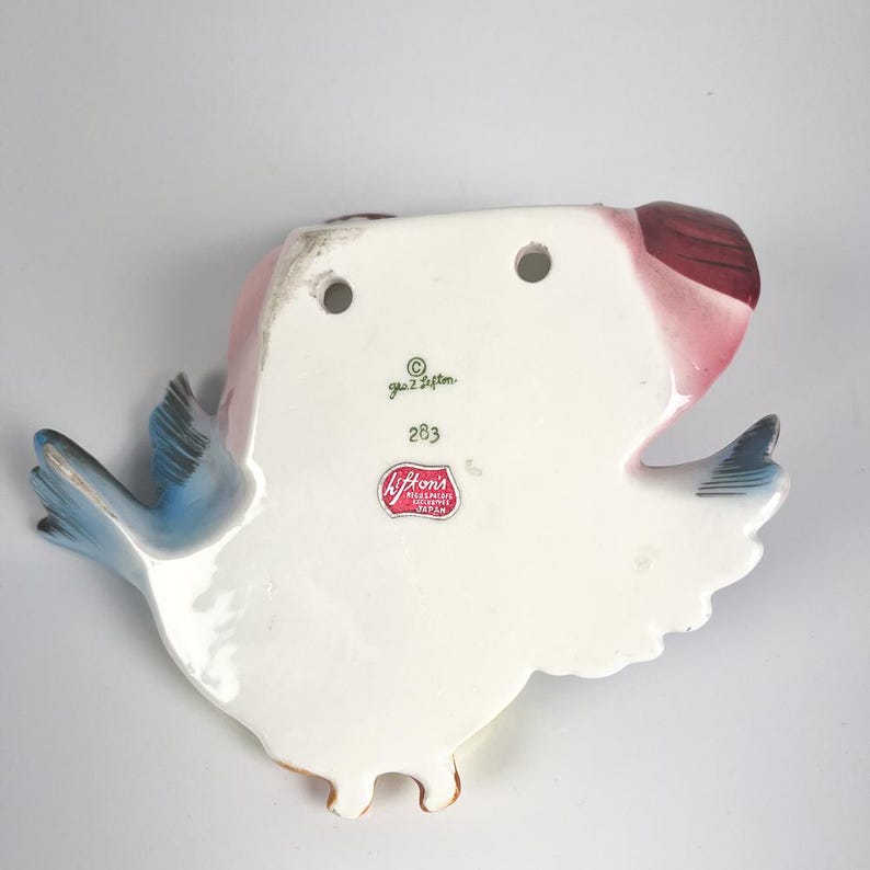 May include: A white ceramic wall pocket shaped like a bird with blue wings and a red tail. The back features two holes for hanging, the text Geo. Z. Lefton, the number 283, and a red sticker label that reads Lefton's Reg. U.S. Pat. Off. Made in Japan.