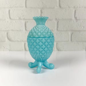 May include: A light blue, translucent glass jar shaped like a pineapple with a removable lid. The jar features a textured, diamond-patterned surface and rests on three curved, decorative feet that extend from the base of the pineapple body.