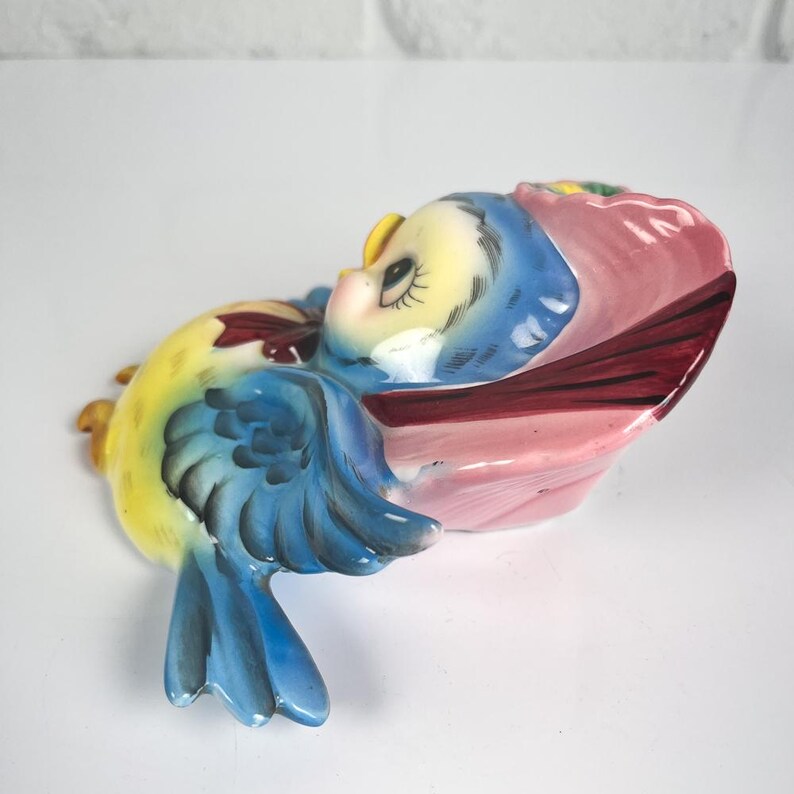 May include: A ceramic wall plaque shaped like a blue and yellow bird wearing a pink bonnet. The bird has a yellow body, blue wings with dark blue detailing, and a pink ruffled hat. The piece features painted facial details including eyes.