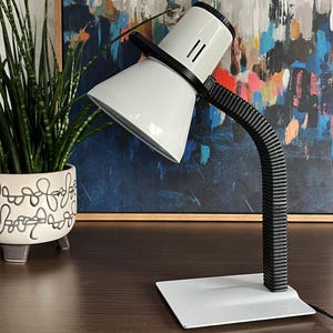 May include: A white desk lamp with a black, flexible neck and base. The lamp has a conical shade and a rectangular base. The lamp is on a dark brown surface. A potted plant is to the left of the lamp.