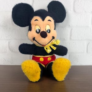 May include: A vintage Mickey Mouse soft toy. The plush has black fur, large round ears, a yellow bow tie, and yellow feet. Mickey is wearing red shorts with yellow buttons. The toy is sitting on a brown surface in front of a white brick wall.