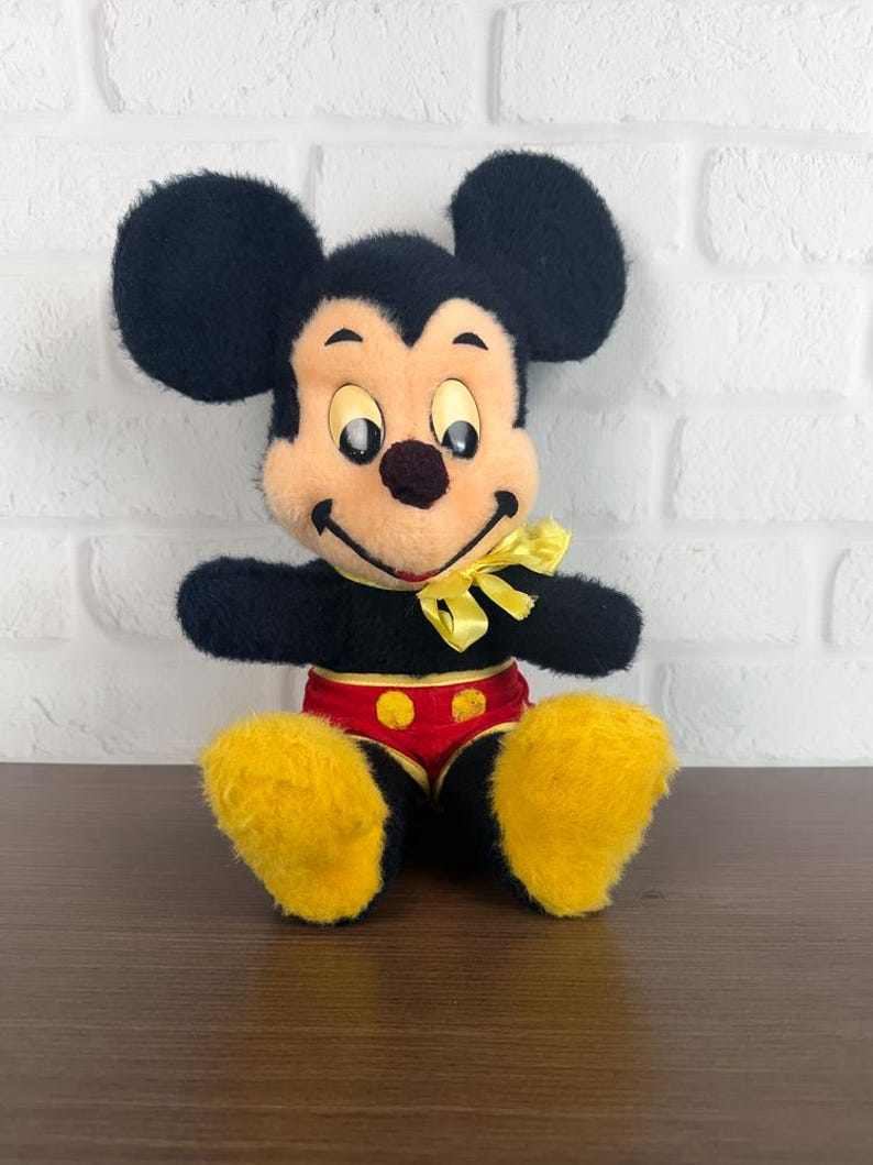 May include: A vintage Mickey Mouse soft toy. The plush has black fur, large round ears, a yellow bow tie, and yellow feet. Mickey is wearing red shorts with yellow buttons. The toy is sitting on a brown surface in front of a white brick wall.