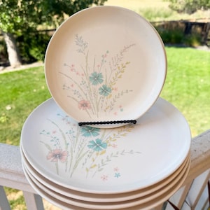 May include: A stack of five round, off-white ceramic plates featuring a delicate floral pattern of blue, pink, and yellow wildflowers. One plate is positioned upright on a black wire stand, while the others are stacked horizontally below it.