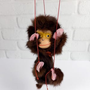 May include: A vintage brown monkey puppet with a yellow face, pink hands and feet, and red strings. The puppet has a furry body and is suspended against a white background. The monkey's expression is cheerful, with a red mouth and a small red bow.