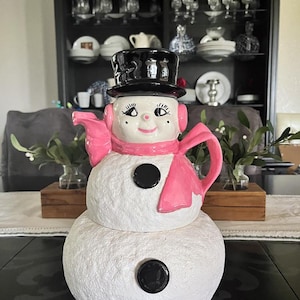 May include: A white snowman-shaped teapot with a black top hat, black buttons, and a pink scarf and ear muffs. The teapot has a smiling face and is likely a holiday or winter-themed decorative item.