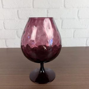 May include: A large, purple-colored glass with a textured surface. The glass has a wide, rounded bowl and a short, dark stem and base. The glass is likely used for serving brandy or cognac.