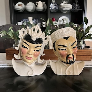 May include: Two ceramic head vases depicting a pair of figures with stylized facial features. The left figure has a cream-colored headpiece and black hair accents, while the right figure wears a cream-colored hat and has a black goatee and mustache.