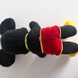 May include: A black plush toy with large ears, wearing red shorts with yellow trim. The toy has yellow feet and a yellow collar. The toy is lying on its back against a white background.
