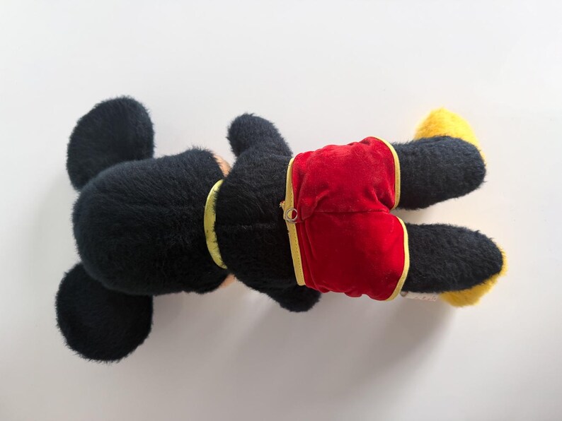 May include: A black plush toy with large ears, wearing red shorts with yellow trim. The toy has yellow feet and a yellow collar. The toy is lying on its back against a white background.