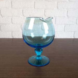 May include: A vintage, turquoise-colored glass creamer with a rounded bowl and a small spout. The creamer sits on a pedestal base and has a matching turquoise stem and foot. The glass is transparent, allowing light to pass through.