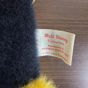 May include: A close-up of a vintage plush toy with black fur and yellow accents. A tag reads "Walt Disney Characters" and "Manufactured by CALIFORNIA STUFFED TOYS". The toy has a red and yellow hat.