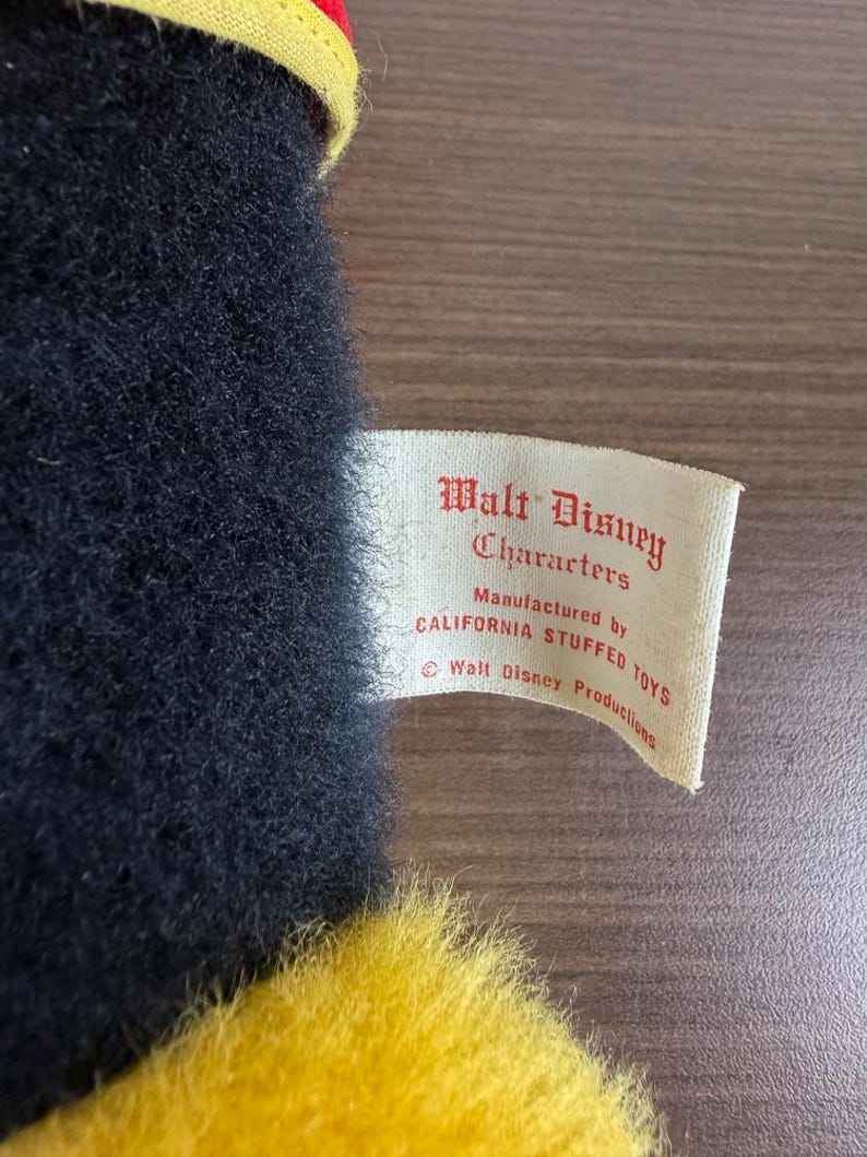May include: A close-up of a vintage plush toy with black fur and yellow accents. A tag reads "Walt Disney Characters" and "Manufactured by CALIFORNIA STUFFED TOYS". The toy has a red and yellow hat.