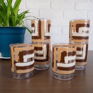 May include: A set of four clear glass tumblers featuring a geometric pattern of horizontal and vertical stripes in shades of dark brown, tan, and cream. Each glass has a cylindrical shape with a slightly recessed base and a smooth rim.