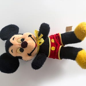 May include: A vintage Mickey Mouse soft toy, with a black body, large ears, and a smiling face with a brown nose. He wears red shorts with yellow buttons and accents, and yellow shoes. A yellow bow is tied around his neck.