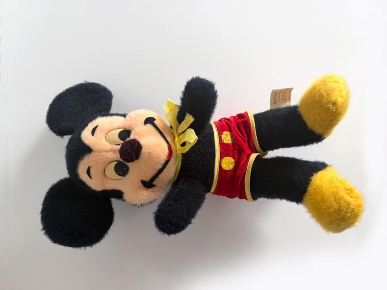 May include: A vintage Mickey Mouse soft toy, with a black body, large ears, and a smiling face with a brown nose. He wears red shorts with yellow buttons and accents, and yellow shoes. A yellow bow is tied around his neck.