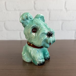 May include: A glossy mint green ceramic dog figurine featuring a textured coat, pointed ears, and a dark brown collar with gold-colored studs. The dog has large dark eyes, a black nose, and a small red tongue visible at the mouth area.