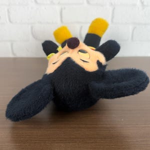 May include: A plush toy of a cartoon character with black fur, yellow ears, and a brown nose. The character has a light orange face and is lying down with its eyes closed. The toy has a soft, fuzzy texture.