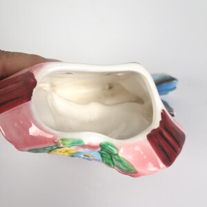 May include: A ceramic wall pocket planter featuring a pink base with hand-painted floral details in yellow, blue, and green. The interior is hollow and white, with dark red accents on the sides and a shaped opening at the top for holding items.