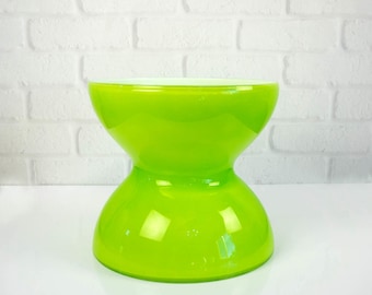 May include: A bright lime green glass stool with a smooth, glossy finish. The stool features a distinct hourglass shape with a wide circular base and a matching wide circular top, connected by a narrower, curved central section.