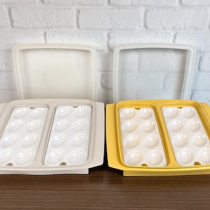 May include: Two plastic egg trays with lids. One tray is beige, the other is yellow. Each tray has two sections, each holding six egg-shaped indentations. The lids are translucent and rectangular.