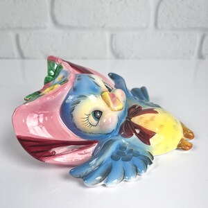 May include: A ceramic wall plaque shaped like a blue bird wearing a pink bonnet with green leaves and yellow flowers. The bird has a yellow chest, blue wings with dark blue detailing, and a brown bow tied around its neck, featuring large eyes.