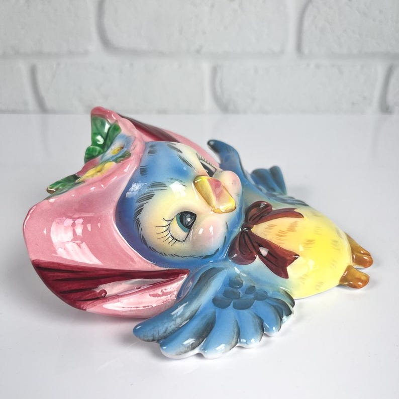 May include: A ceramic wall plaque shaped like a blue bird wearing a pink bonnet with green leaves and yellow flowers. The bird has a yellow chest, blue wings with dark blue detailing, and a brown bow tied around its neck, featuring large eyes.
