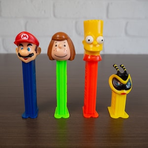 May include: Five colorful plastic toy figures with different characters, including Mario, a girl with brown hair, a yellow figure with a yellow and orange striped shirt, and a yellow bee figure. The figures are all on a brown wooden surface.
