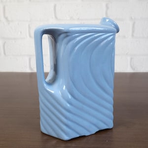 May include: A light blue ceramic pitcher with a handle and a spout. The pitcher has a rectangular shape with a textured, wave-like design on the sides. The pitcher is sitting on a brown surface.
