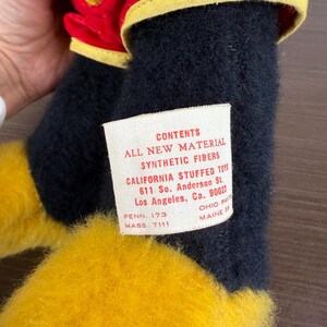 May include: A close-up of a stuffed toy with black, yellow, and red fabric. The toy has a tag that reads "CONTENTS ALL NEW MATERIAL SYNTHETIC FIBERS CALIFORNIA STUFFED TOYS 611 So. Anderson St. Los Angeles, Ca. 90023".