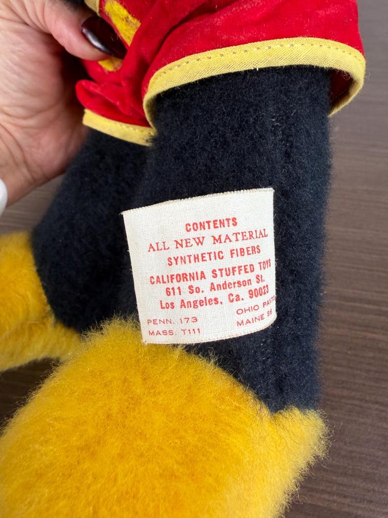 May include: A close-up of a stuffed toy with black, yellow, and red fabric. The toy has a tag that reads "CONTENTS ALL NEW MATERIAL SYNTHETIC FIBERS CALIFORNIA STUFFED TOYS 611 So. Anderson St. Los Angeles, Ca. 90023".