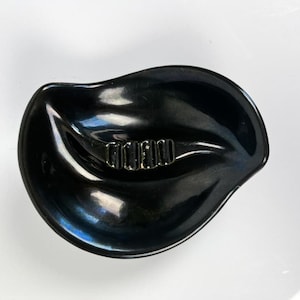 May include: A glossy black ashtray with a unique, organic shape. The ashtray features a central rest area with three slots for cigarettes. The design is sleek and modern, with a reflective surface.