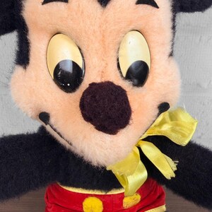 May include: A close-up of a vintage Mickey Mouse soft toy. The toy has a black body, a peach-coloured face, and large, oval eyes. Mickey is wearing red shorts with yellow buttons and a yellow bow tie. A small tag is visible.