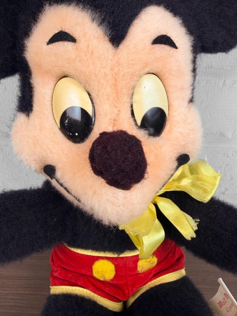 May include: A close-up of a vintage Mickey Mouse soft toy. The toy has a black body, a peach-coloured face, and large, oval eyes. Mickey is wearing red shorts with yellow buttons and a yellow bow tie. A small tag is visible.