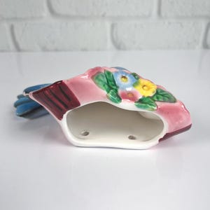 May include: A ceramic wall pocket shaped like a bird, featuring a pink body with a blue tail and wing. The top section is decorated with a cluster of small, colorful flowers in yellow, blue, and pink with green leaves. The interior is hollow and white.