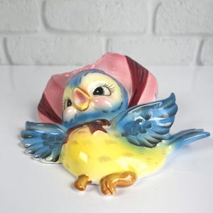 May include: A ceramic wall pocket shaped like a blue bird with a yellow belly and a pink bonnet. The bird has large eyes with long lashes, a yellow beak, and outstretched wings. A small brown bow is painted at the neck of the bird figure.
