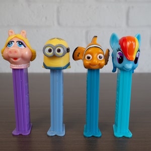 May include: Five colorful character-themed plastic toy dispensers. The dispensers are purple, blue, and light blue. The characters are Miss Piggy, a Minion, Nemo, and a My Little Pony.