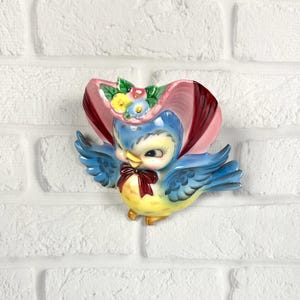 May include: A ceramic wall plaque shaped like a blue bird wearing a pink bonnet with small yellow, blue, and pink flowers. The bird has a yellow chest, blue wings, and a dark red bow tie around its neck. The finish is glossy and colorful.