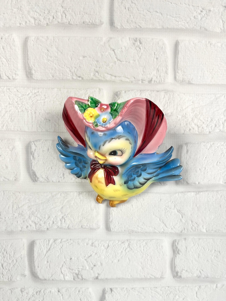 May include: A ceramic wall plaque shaped like a blue bird wearing a pink bonnet with small yellow, blue, and pink flowers. The bird has a yellow chest, blue wings, and a dark red bow tie around its neck. The finish is glossy and colorful.