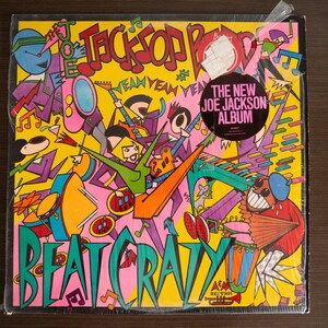 May include: A colorful vinyl record cover with a cartoon illustration of people playing music. The cover is yellow, pink, green, blue, and red. The title of the album is "Beat Crazy" and the text "The New Joe Jackson Album" is in a circle on the cover.