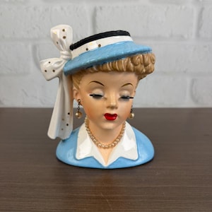 May include: A ceramic head vase depicting a person with brown hair, wearing a blue hat with a white bow featuring gold dots, a blue collared shirt, a pearl necklace, and dangling pearl earrings with red lips and closed eyes.