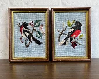 May include: Two framed needlepoint pictures of birds on branches. The birds are black, white, and red. The branches have green leaves and red berries.