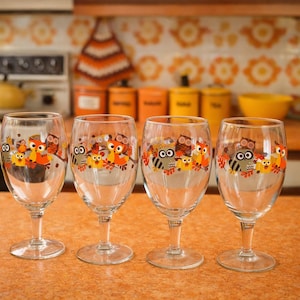 May include: Four clear wine glasses with a colorful owl design around the rim. The owls are in shades of orange, yellow, and brown. The glasses are on an orange countertop, with a retro kitchen in the background.