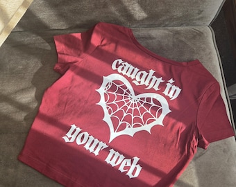 May include: A maroon short-sleeved t-shirt featuring a white graphic of a heart-shaped spider web. The text "caught in" is printed above the heart, and "your web" is printed below the heart in a bold, gothic-style white font.