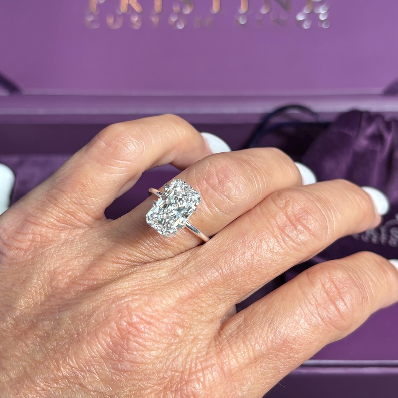 May include: A radiant-cut diamond ring with a silver band. The large, rectangular diamond sparkles brightly. The ring is displayed on a hand with a purple background. The word "KRISTINA" is visible in the background.