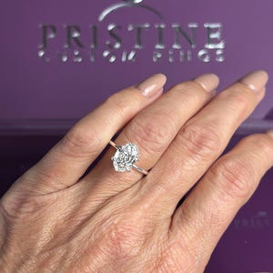 K&ouml;nnte beinhalten: Ein silberfarbener Ring mit einem gro&szlig;en, oval geschliffenen, klaren facettierten Edelstein in einer einfachen Metallschiene. Der Ring befindet sich vor einer violetten Schachtel mit dem silbernen Text PRISTINE CUSTOM RINGS und einem Ring-Symbol.