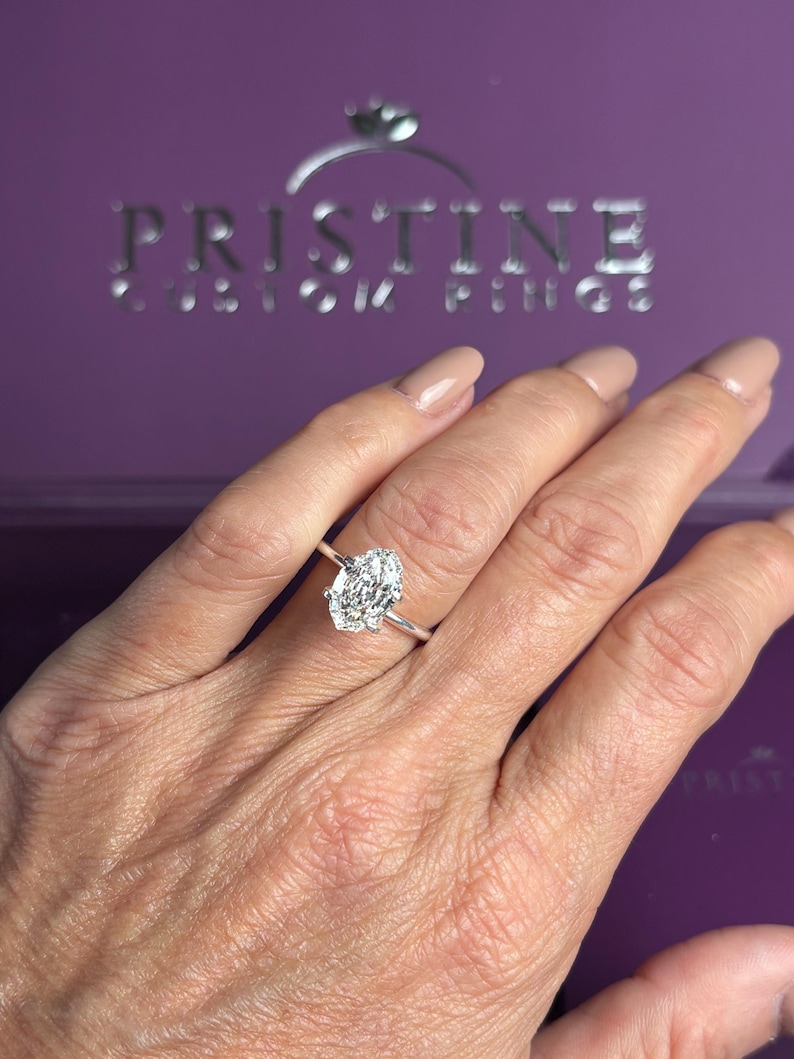 K&ouml;nnte beinhalten: Ein silberfarbener Ring mit einem gro&szlig;en, oval geschliffenen, klaren facettierten Edelstein in einer einfachen Metallschiene. Der Ring befindet sich vor einer violetten Schachtel mit dem silbernen Text PRISTINE CUSTOM RINGS und einem Ring-Symbol.