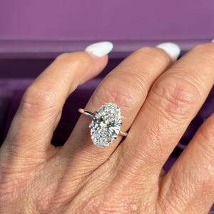 May include: A silver engagement ring with a large oval-cut diamond center stone and a halo of smaller diamonds. The ring is on a person's finger, with a purple box in the background. The word "PRISTINE" is visible on the box.
