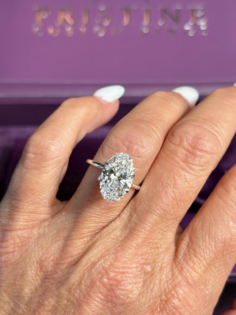 May include: A silver engagement ring with a large oval-cut diamond center stone and a halo of smaller diamonds. The ring is on a person's finger, with a purple box in the background. The word "PRISTINE" is visible on the box.