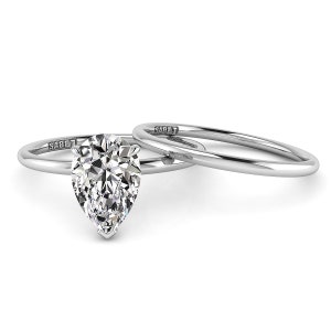 Puede incluir: Un anillo de compromiso de diamante de forma pera engastado en oro blanco con una banda de boda a juego. El anillo est&aacute; grabado con la palabra "SABET" en la banda.